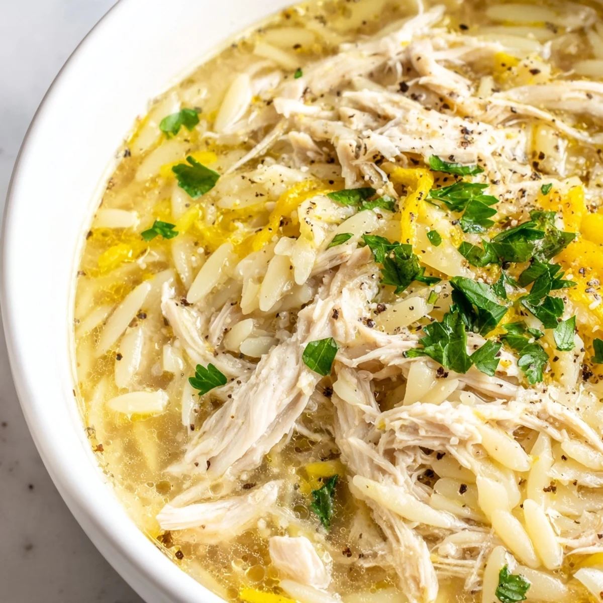 Golden Lemon Pepper Chicken Orzo Soup steams in a white bowl, with tender shredded chicken, carrots, and parsley.